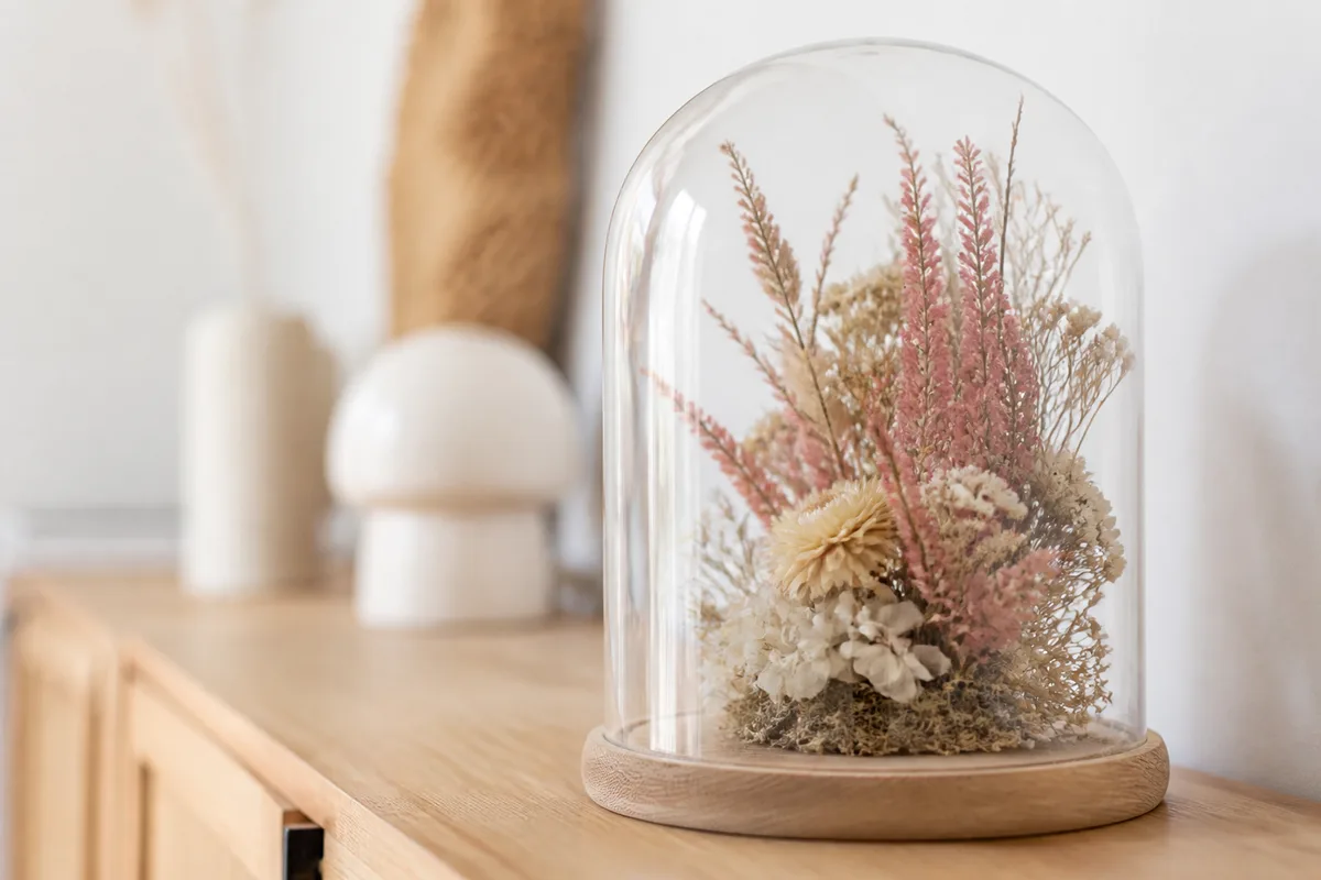 Cloche en verre sur socle en bois clair abritant une composition de fleurs séchées beiges et roses, posée sur une console en bois près d’un mur blanc, lumière naturelle latérale douce