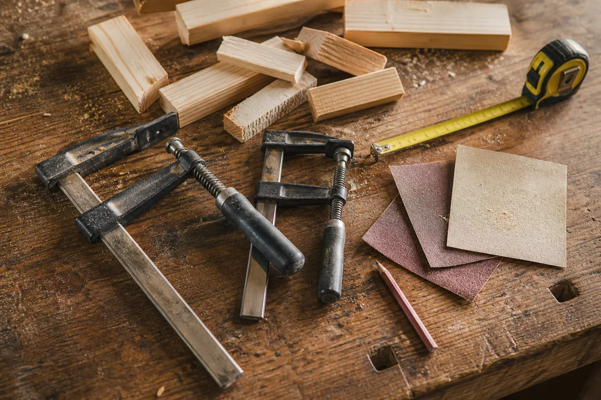 Établi d&rsquo;atelier avec chutes de tasseaux en bois clair, serre-joints métalliques, mètre ruban et papier de verre, lumière latérale douce